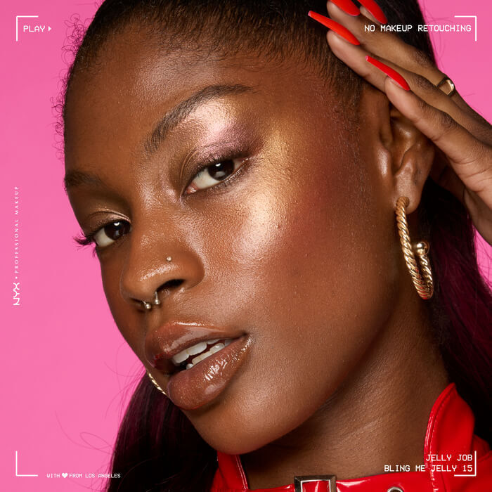 Close-up of a model with glossy lips, wearing red-toned eyeshadow and holding a red NYX Jelly Job lip gloss. Background is pink. Text reads: "Jelly Job - Orange Ya Jelly? 13".-2