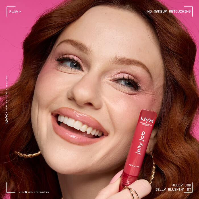 Close-up of a model with glossy lips, wearing red-toned eyeshadow and holding a red NYX Jelly Job lip gloss. Background is pink. Text reads: "Jelly Job - Orange Ya Jelly? 13".-1