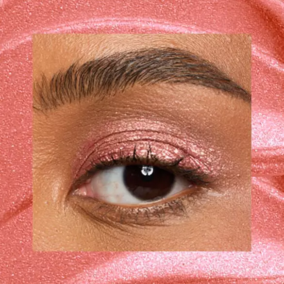 Close-up of an eye area with NYX Professional Makeup Lid Lingerie Cream Eye Pigment in 09 Cheeky Coral, shown on a coral shimmer textured background.
