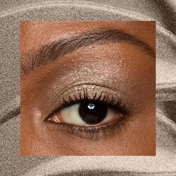 Close-up of an eye area with NYX Professional Makeup Lid Lingerie Cream Eye Pigment in 07 Granite Grind, shown on a brown shimmer textured background.