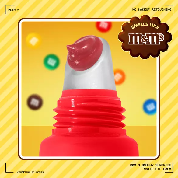 Close-up of NYX Professional Makeup M&M’S Smushy Surprize Matte Lip Balm with a red applicator tip, set against a yellow background with colorful M&M’S candies. Text highlights M&M’S scent and no makeup retouching.