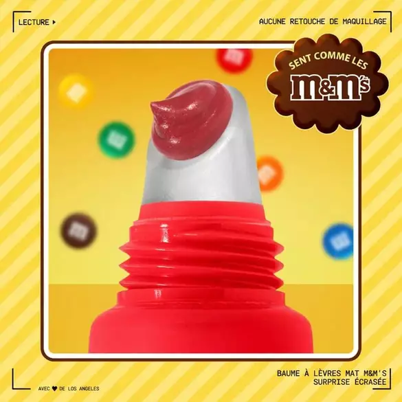 Close-up of NYX Professional Makeup M&M’S Smushy Surprize Matte Lip Balm with a red applicator tip, set against a yellow background with colorful M&M’S candies. Text highlights M&M’S scent and no makeup retouching.
